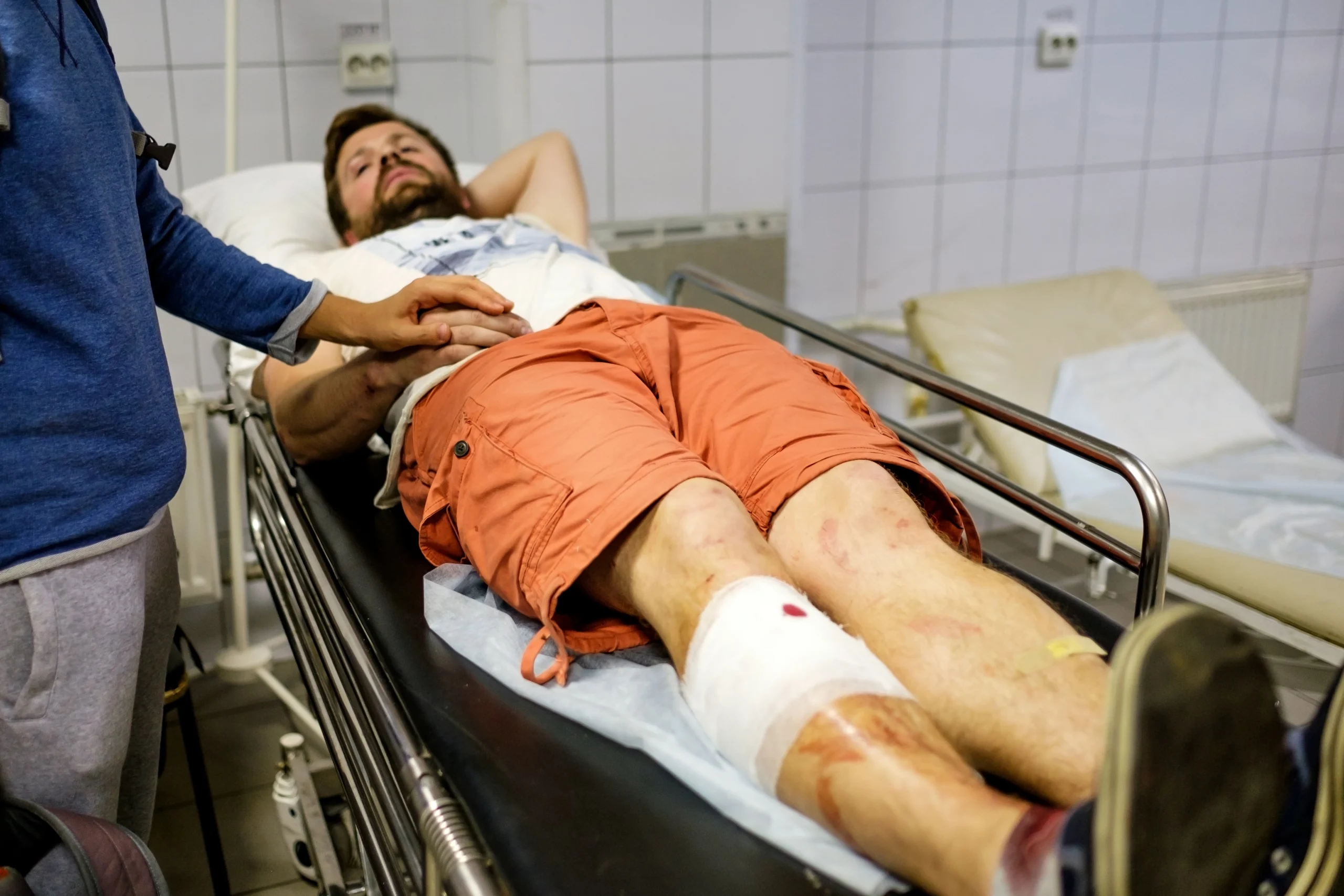 A man with a leg injury lies on a hospital bed, wearing an orange shirt. His bandaged leg is visible as another person holds his hand, offering support.